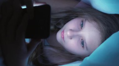 Dikey video. Çocuk bağımlılığı. Çevrimiçi gece. Odaklanmış, takıntılı bir kız. Akıllı telefonuyla battaniyenin altına saklanıyor. Evde yatarken içeriği izliyor..