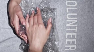 Dikey video. Plastik küçültme. Sıfır kayıp. Kırışık şişeleri olan gönüllü bir kadın gri arka planda çöpleri geri dönüştürüyor..