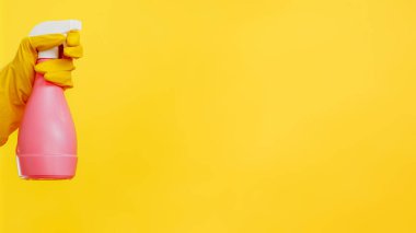 Ev işi aletleri. Hijyen ürünleri. Deterjan ekipmanı. Elinde pembe temizleyici sprey şişesi olan, sarı arka planda izole edilmiş koruyucu eldivenli bir hademe..
