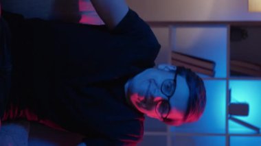 Dikey video. El sıkışıyoruz. Kafası karışmış bir adam. Meme ifadesi. Gözlüklü cahil komik adam, el sıkışıyor, karanlık neon ışıklı evin içinde oturuyor..