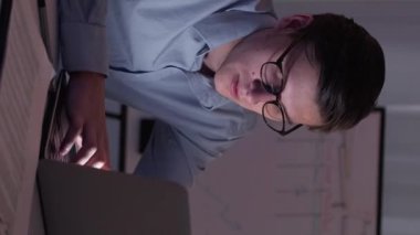 Dikey video. Erkek işçi. Zihinsel aşırı yüklenme. İş raporu. Yorgun akıllı adam ofiste bilgisayarla çok çalışıyor. Oturma odasında..