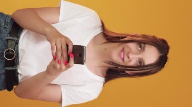 Dikey video. Romantik akıllı telefon sohbeti. Kurye ilanı. Çevrimiçi eğlenceli iletişim. Mutlu flörtçü düşünceli kadın turuncu arka planda hücre öpücüklerini yazıyor..