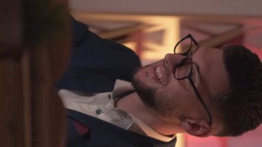 Dikey video. Çevrimiçi iletişim. Gülümseyen adam. Ev ödevi. İçi karanlık oda kitaplarıyla dolu dizüstü bilgisayarda oturan mutlu zeki adam..