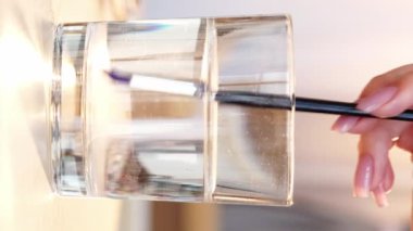 Dikey video. Suluboya resim. Çizim işlemi. Kadın ressamın eli, masasında mavi renkli su bardağıyla ıslak boya fırçasıyla yakın plan..