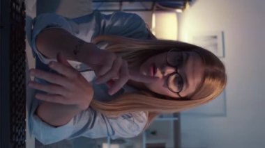 Dikey video. İnternetteki konferans. Zeki kadın. Mesafe çalışması. Gözlüklü, esinlenilmiş, karanlık ışık odasındaki masasında konuşan güzel bir bayan..