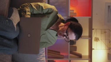 Dikey video. Laptop yardımı. Bilgisayar açıklıyor. Anne not defteri programını gösteren genç bir kız. İnternet. Akşam kanepede oturuyor..
