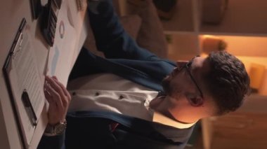 Dikey video. Muhasebe süreci. Erkek bir uzman. Finansal hesaplamalar. Yoğunlaşmış adam notlar alıyor. Belgeler üzerinde çalışıyor. Karanlık ışık odasının iç tarafında oturuyor..