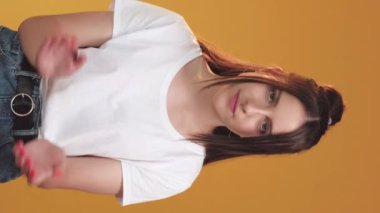 Dikey video. Alaycı bir ifade. Şüpheci duygular. Hayal kırıklığına uğramış, el çırpan şüpheli bir kadın. Turuncu arka planda izole edilmiş el kol işareti var..