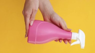 Dikey video. Temizlik spreyi. Ev hijyeni. Kadın elleri, sarı arka planda izole edilmiş yüzey deterjan sıvısıyla pembe şişe reklamı yapıyor..