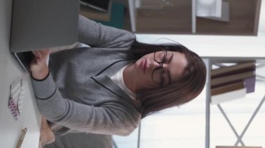 Dikey video. Yorgun iş. Yorgun bir kadın. Uzaktan kumanda. Hasta kadın burnuna masaj yapıyor. Oturma masası ışık odasının iç tarafında dizüstü bilgisayar açıyor..