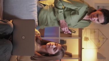 Dikey video. Bilgisayar açıklıyor. Laptop yardımı. Genç kızı gösteren bir kadın dizüstü bilgisayar programı kullanıyor. İnternette akşam kanepede oturuyor..