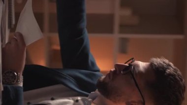 Dikey video. Bilgileri. Endişeli adam. İş başarısız oldu. Gerekli belgeleri bulmak için kendini çaresiz hisseden meşgul bir adam karanlık odanın içinde masa başında oturuyor..