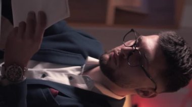 Dikey video. İş projesi. Meşgul adam. Soruşturma işi. Akıllı, konsantre bir adam masa lambası odasının içindeki belgeleri inceliyor..