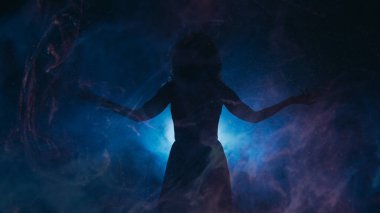 Spotlight silhouette. Spiritual energy. Woman with spread arms in haze shadow blue projector glowing particles fog dark background copy space. clipart