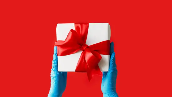 Christmas present. Gift delivery. Unrecognizable human hands in protective gloves holding wrapped box quarantine hygiene measures isolated on red free space background.