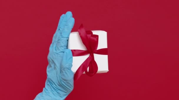 バーティカルビデオ クリスマスサプライズ パンデミック配達 赤いコピースペースの背景に隔離されたリボン弓が付いている白い箱で示す青い医学の手袋の手 — ストック動画