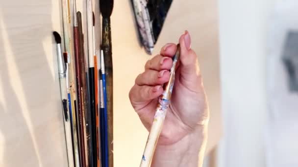バーティカルビデオ アートツール クリエイティブアクセサリー アートワークのテーブルスタジオの職場で描くためのペイントブラシを選択する女性画家の手のクローズアップ — ストック動画