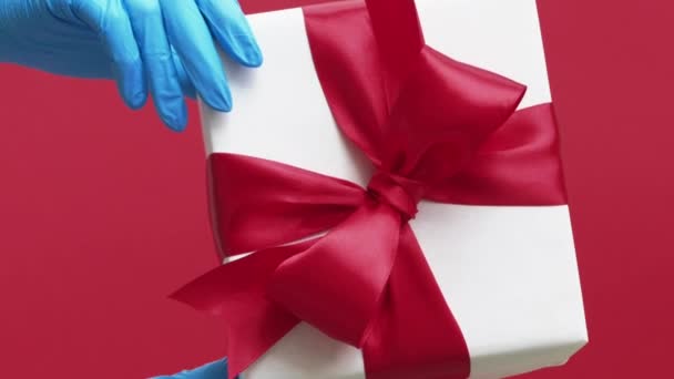 バーティカルビデオ パンデミックギフト クォンタインプレゼント 赤い背景に隔離されたリボン弓の白い包まれた箱を示す青い保護手袋の手 — ストック動画