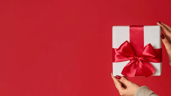Christmas surprise. Gift delivery. Unrecognizable female hands showing white wrapped present in box with ribbon bow isolated on red copy space background.