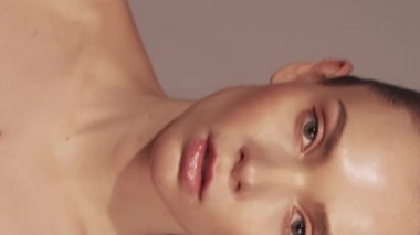 Dikey video. Moda modeli yaşam tarzı. Güzellik fotoğrafı çekimi. Çıplak makyajlı, kendine güvenen, parlak yüzlü, çıplak omuzlu, flaşlı, bej arkaplanlı bir kadın..