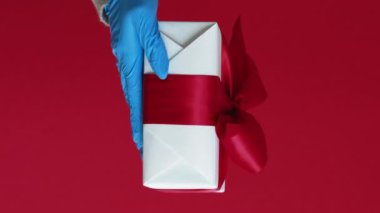 Dikey video. Doğum günü hediyesi. Coronavirus mevcut. Kırmızı fotokopi arkaplanında izole edilmiş, koruyucu eldivenli, tanınmayan insan eli..