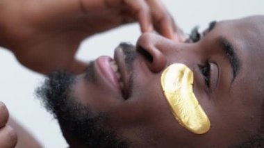 Dikey video. Erkek yüz bakımı. Cilt tazeleme. Sabah rutiniyle yakışıklı, mutlu bir adam. Parlak yüzünde altın rengi kolajen jel göz bandı..