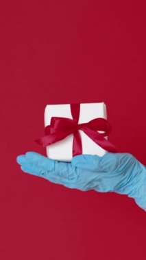 Dikey video. Noel sürprizi. Pandemik doğum. Mavi tıbbi eldiven, kırmızı fotokopi arkaplanda izole edilmiş kurdele yayı ile beyaz kutuda görünüyor..