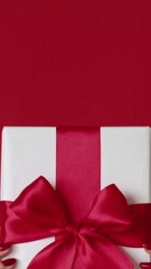 Dikey video. Tatil hediyesi. Hediye teslimatı. Kırmızı boş arkaplanda izole edilmiş beyaz kurdele yayı özel teklifi gösteren kadın elleri.