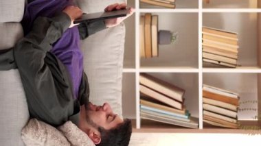 Dikey video. Cihaz dinlendirici. İnternet eğlencesi. Hafta sonu evde. Dijital tablette sörf yapan mutlu bir adam oturma odasında rahat bir kanepenin keyfini çıkarıyor..