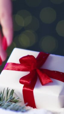 Dikey video. Mutlu noeller. Hediye sürprizi. Xmas dekorasyonu. Kadın el ele tutuşup kırmızı fiyonklu hediye kutusunu kışın kar çamı kompozisyonu Bokeh 'in arka planına koyuyor..