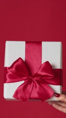 Dikey video. Yeni yıl sürprizi. Noel hediyesi. Kırmızı boş arkaplanda izole edilmiş kurdele yayı ve beyaz paketi gösteren iki kadın eli..