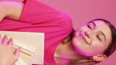 Dikey video. Edebiyat günü. Kitap festivali. Okuma hobisi. Pasifiğin pembe arka planında çok satan roman yığınıyla dans eden mutlu tatlı kadın..