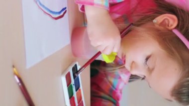 Dikey video. Çocuk ressam. Yaratıcı bir eğlence. Yetenekli çocuklar için okuldaki masada suluboya ve boya fırçasıyla gökkuşağı resmi yapan küçük bir kız..