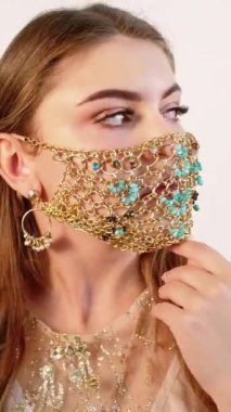 Dikey video. Karantina güzelliği. Salgın mücevherler. Şehvetli esmer kadın altın zincirli poz veriyor. Yüz maskesi, açık arkaplanda izole edilmiş lüks kıyafete uyuyor..