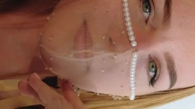 Dikey video. Şenlikli parti bakışı. Pandemik moda. Pırıl pırıl parıldayan inci desenli boncuklar takmış zarif bir kadın hafif arka planda kameraya nazikçe poz veriyor..