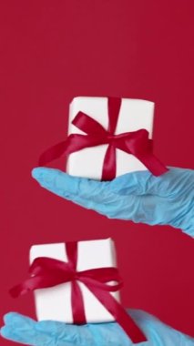 Dikey video. Noel hediyesi. Yeni yıl salgını. Mavi eldivenli kadın eller kırmızı boş alanda izole edilmiş hediyelerle dolu iki kutu tutuyor..