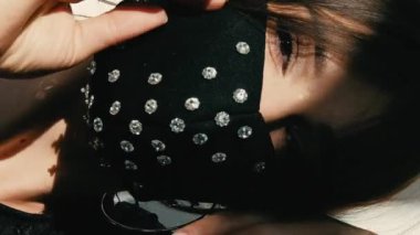 Dikey video. Karantina modası. Şık bir parti havası. Koyu renk desenli paillette elbisesi ve kristal maskeli kadın dans eden güneş ışığı gölgelerinin keyfini çıkarıyor..