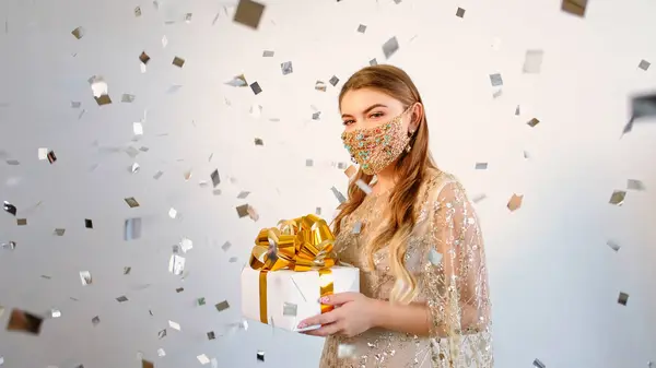 Holiday surprise. Pandemic party. Happy woman in DIY gold chain face mask matching dress holding gift box in confetti rain on light copy space background.