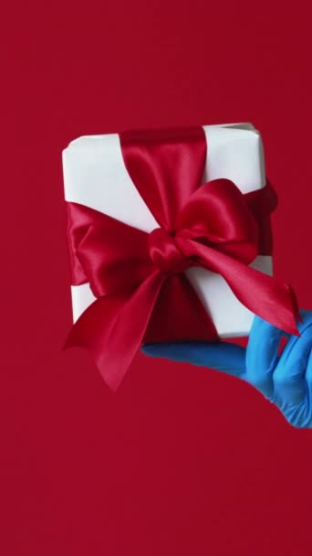 バーティカルビデオ プレゼント配信 ホリデーギフト 赤い背景に隔離されたジェスチャーのような包まれた箱の親指を握る保護手の認識できない人間の手 — ストック動画