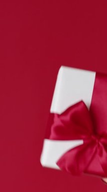 Dikey video. Noel hediyesi. Kış tatili sürprizi. Kırmızı boş arkaplanda izole edilmiş parlak kurdele yayı ile beyaz hediye kutusu gösteren kadın elleri.