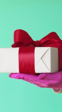 Dikey video. Doğum günü hediyesi. Hediye sunuyorum. Koruyucu eldivenler içinde tanınmayan insan eli mavi boş bir alanda izole edilmiş kutu hijyeni tutuyor..