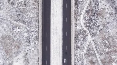 Dikey video. Hava karlı görünüyor. Kırsal kış yolu. Beyaz donmuş tarlalar, asfalt pist ve geçen arabalarla trafik yolu bulutlu hava aracı..