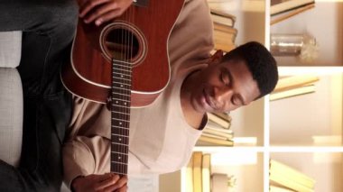 Dikey video. Gitar alıştırması. Müzisyen eğlencesi. Yaratıcı hobi. Yetenekli, neşeli bir adam evdeki oturma odasındaki kanepede telli çalgılarla şarkı söylüyor..