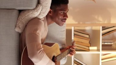 Dikey video. Gitar dinlendirici. Müzikal eğlence. Ev eğlencesi. Sakin, rüya gibi bir adam akustik telli enstrümanla şarkı söylüyor. Oturma odasındaki hafta sonu kanepesinin keyfini çıkarıyor..