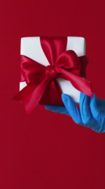 Dikey video. Hediye teslimatı. Hediye. Koruyucu eldivenli, kutunun başparmağını kaldıran tanınmamış insan eli kırmızı arka planda izole edilmiş..