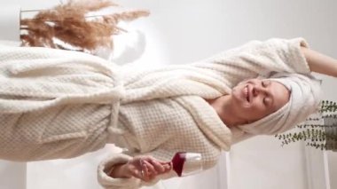 Dikey video. Sabah keyfi. Spa içkisi. Hafta sonu eğlencesi. Evde kırmızı şarap kadehiyle dans eden bornozlu rahat bir kadın..