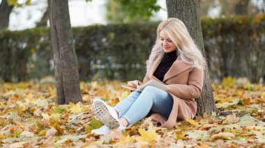 Okumaktan zevk alıyorum. İlham verici bir kadın. Güzel sonbahar. Gülümseyen kadın, elinde ilginç bir kitapla parkta yere yaslanıyor..