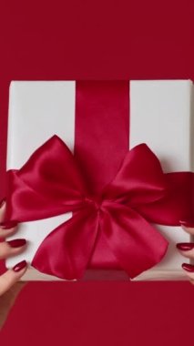 Dikey video. Yeni yıl hediyesi. Noel hediyesi. Kırmızı arka planda izole edilmiş bayanların elleri sarılı beyaz kutu ve kurdele yayı bayramı kutlaması..