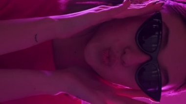 Dikey video. Şık gözlükler. Hippi kız. Neon ışıklı portre. Pembe arka planda kendine güvenen havalı kadın yüzü..