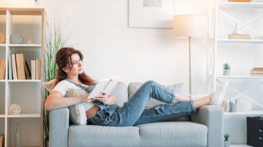 Okumaktan zevk alıyorum. İlham verici bir kadın. Evde rahatla. Gülümseyen orta yaşlı bayan, aydınlık odanın içinde elinde ilginç bir kitapla kanepede yatıyor..